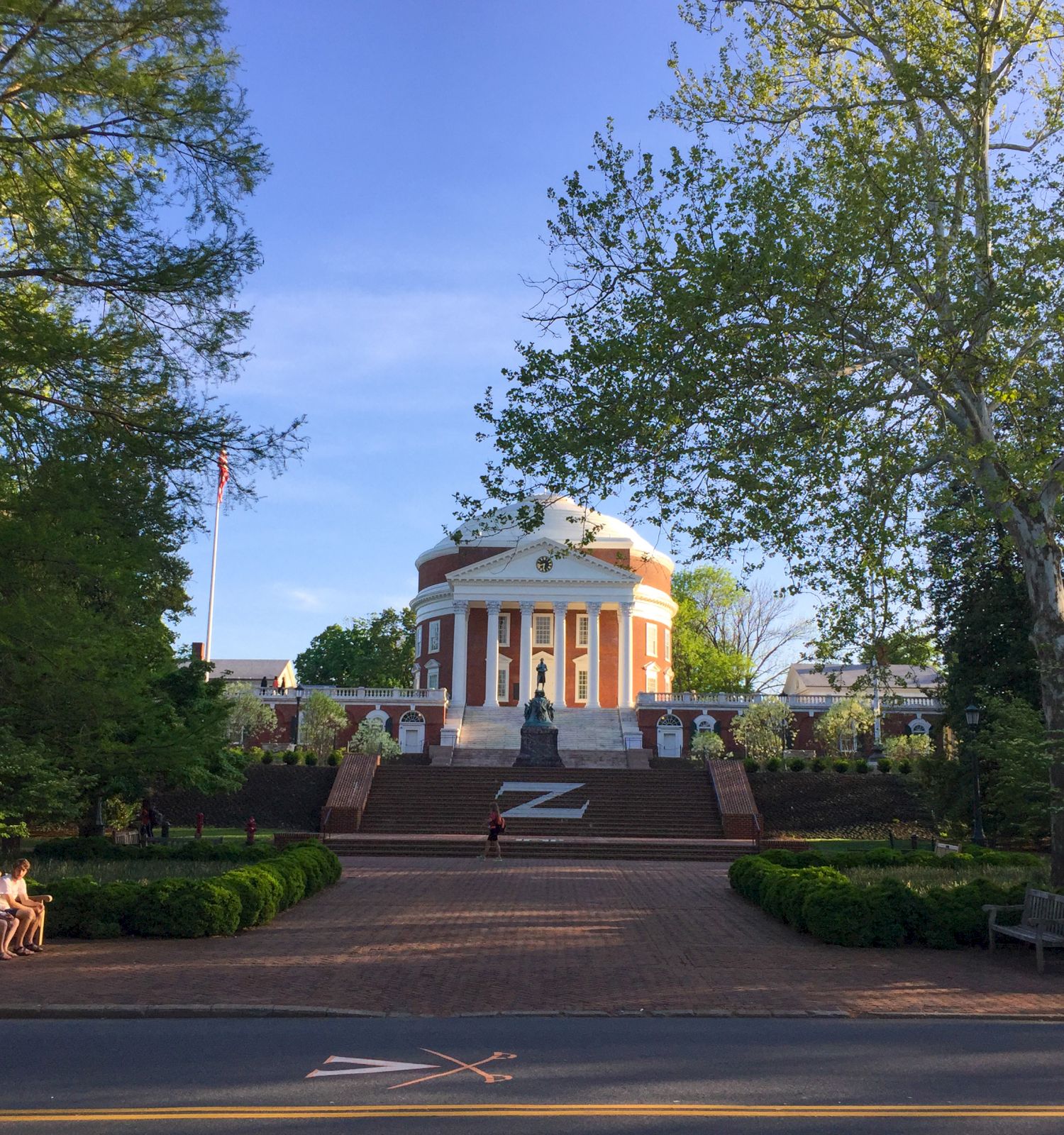 A sunny view of a grand red-and-white neoclassical building with columns and stairs, surrounded by lush green trees and flags.