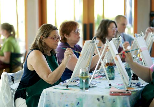 People sit at tables painting with easels and brushes in a classroom/workshop.