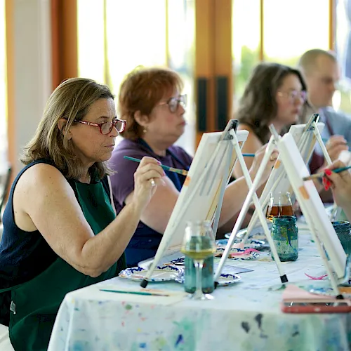 People sit at tables painting with easels and brushes in a classroom/workshop.