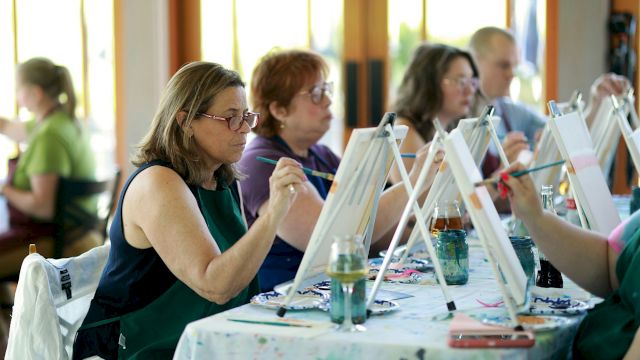 People sit at tables painting with easels and brushes in a classroom/workshop.