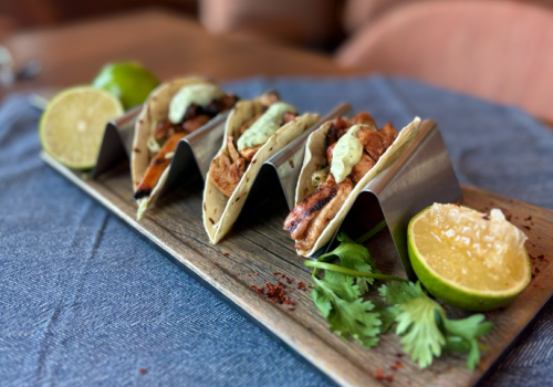 Three tacos on a wooden board, topped with sauce and herbs, with lime wedges on a blue cloth.