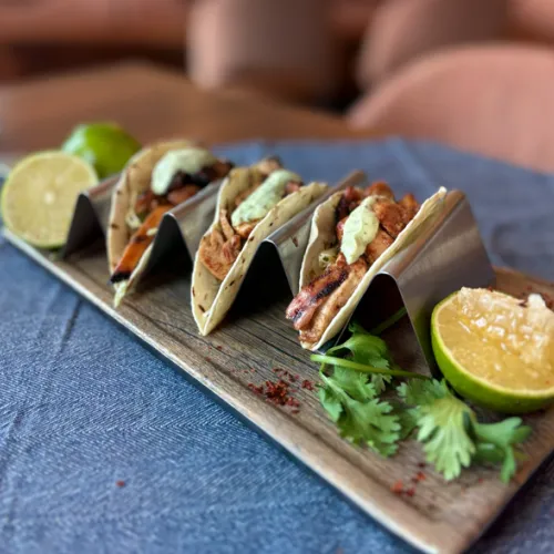 Three tacos on a wooden board, topped with sauce and herbs, with lime wedges on a blue cloth.