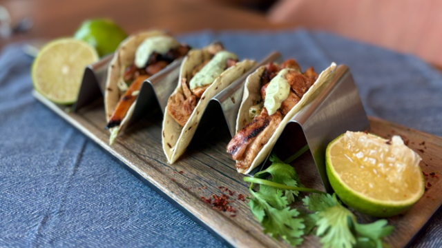 Three tacos on a wooden board, topped with sauce and herbs, with lime wedges on a blue cloth.
