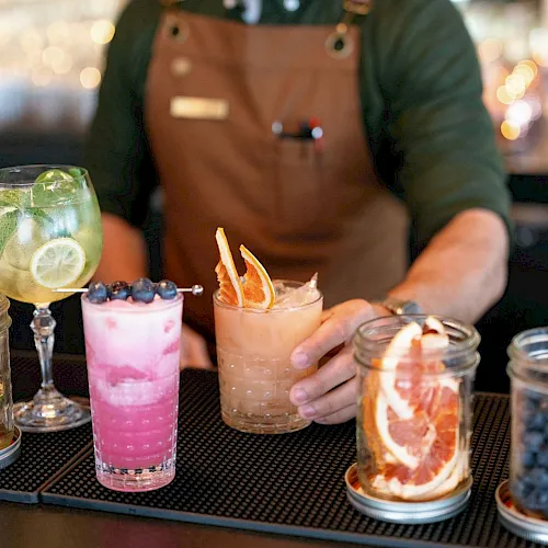 A bartender in an apron smiles while serving colorful cocktails&mdash;pink berry, pale orange, and a lime drink with blueberries.