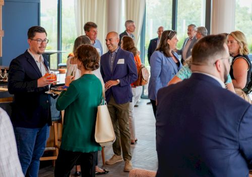 A business networking event with people chatting and holding drinks in a bright room with large windows.