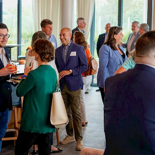 A business networking event with people chatting and holding drinks in a bright room with large windows.