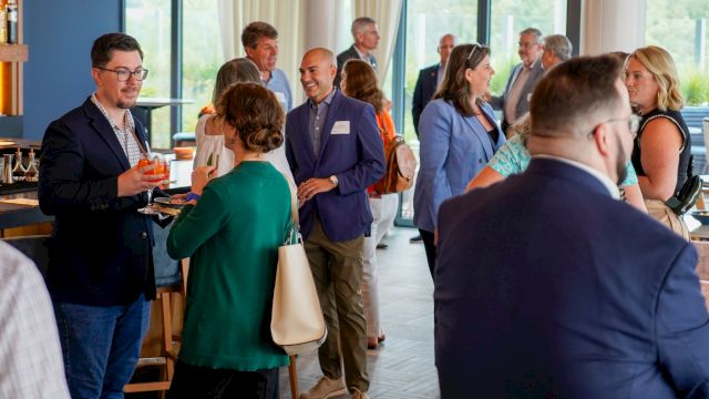 A business networking event with people chatting and holding drinks in a bright room with large windows.