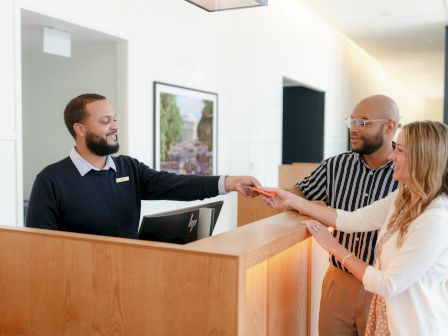 In a bright office, two men and a woman exchange a red document at a reception desk.