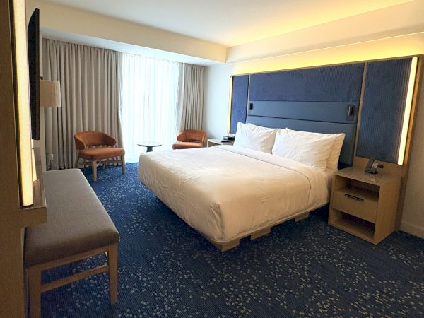 A hotel room with a king bed, bedside tables, lamps, curtains, and a couple of chairs.