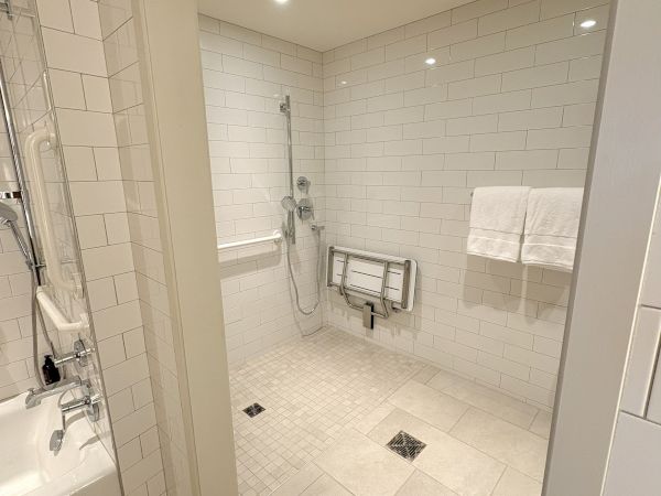 Bright tiled walk-in shower with grab bars, handheld shower, towel rails, and a folding seat in a modern bathroom.