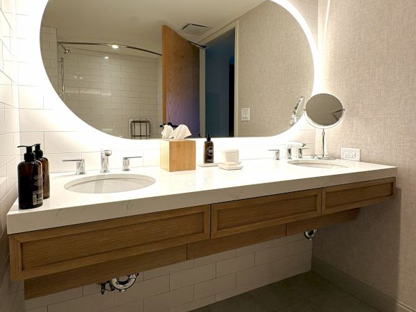A bright modern bathroom vanity with a large round mirror, double sinks, soap bottles, towels, and a small magnifying mirror.