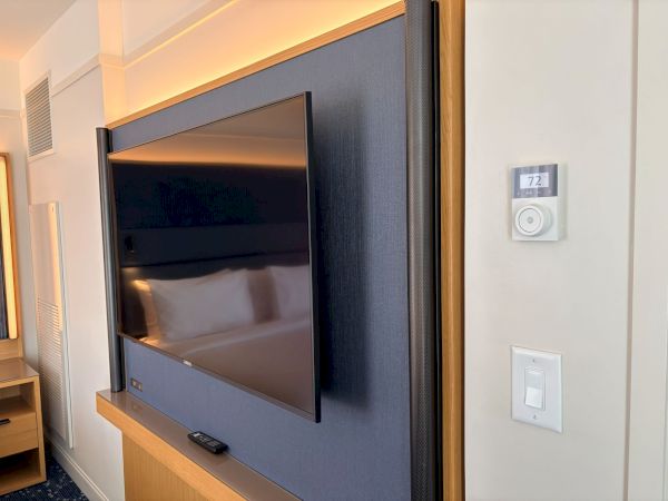 Hotel room wall with a flat-screen TV, warm LED backlight, and thermostat reading 72&deg;F beside a light switch.