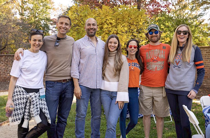 Seven friends smile for a fall outdoor group photo, wearing casual tees and sunglasses beside a brick wall.