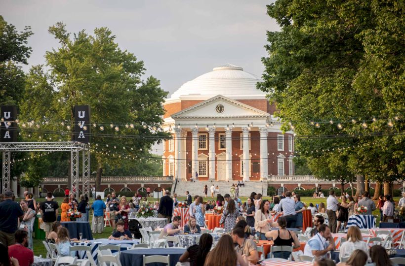 Crowd dining outdoors in front of a white-domed brick building with columns and clock as string lights hang overhead.