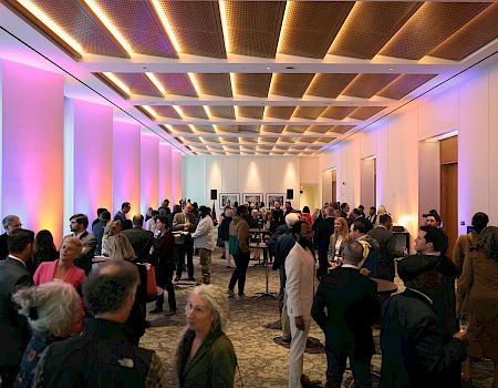 A conference networking event in a modern hall with attendees, cocktail tables, and framed displays under colorful lighting.