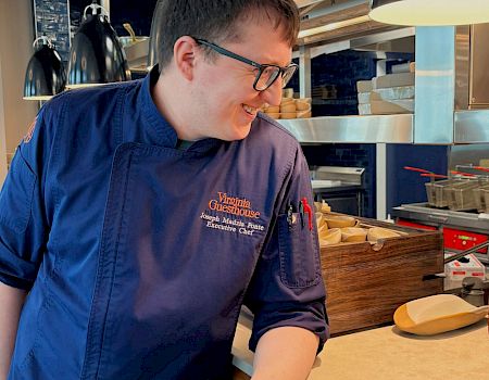 A chef in a Virginia Guesthouse jacket in a commercial kitchen, smiling while plating food.