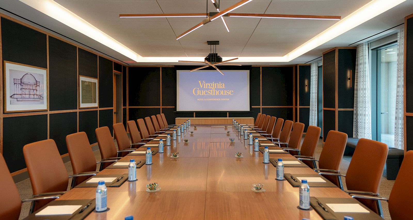 A modern conference room set for a meeting, with &ldquo;Virginia Guesthouse&rdquo; on a screen and rows of chairs around a long table.
