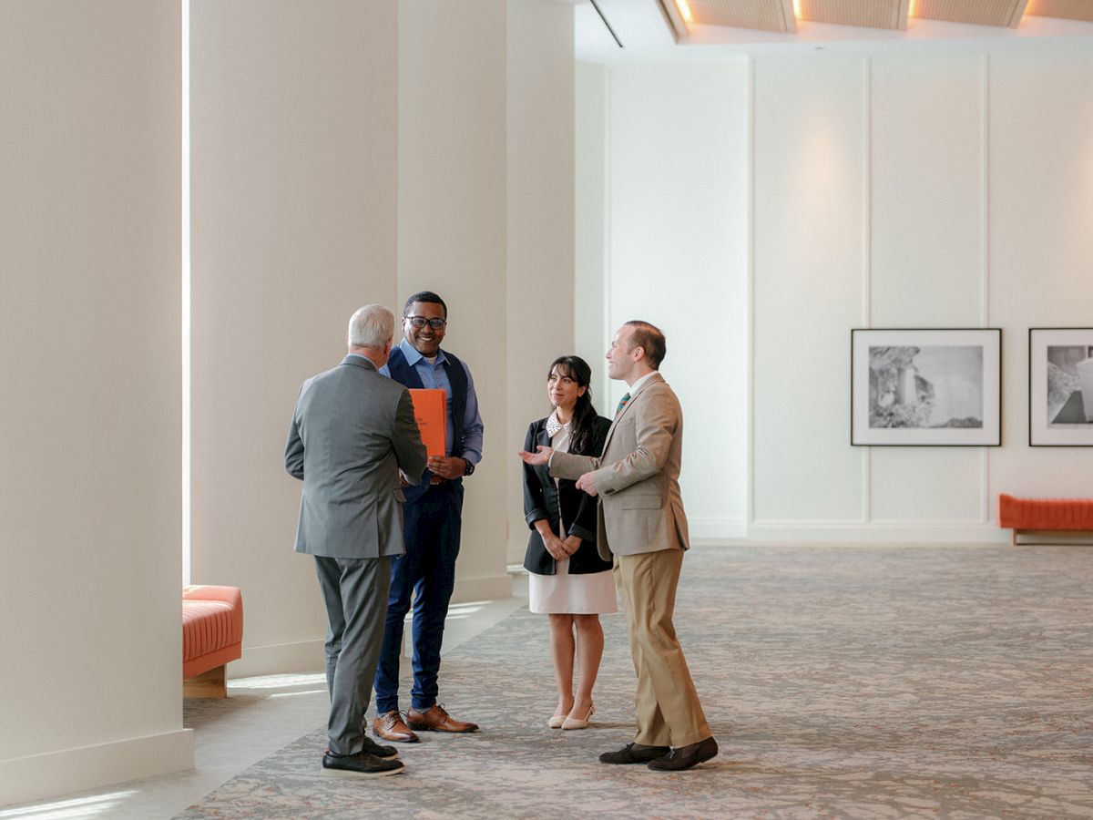 A few business professionals chat and exchange a folder in a bright office lounge with framed photos on the wall.