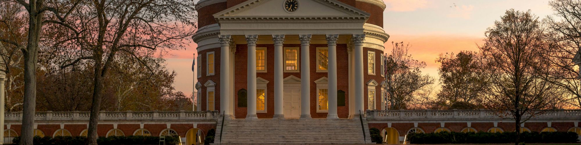 A domed brick neoclassical building with tall columns and a clock faces the sunset behind leafless trees, in a campus setting.