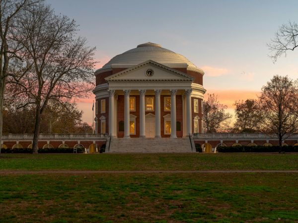 A grand brick rotunda with tall white columns and a clock faces the sunset, framed by leafless trees and a lawn.