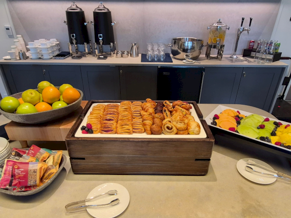 A hotel breakfast buffet with pastries, sliced fruit, and hot-beverage dispensers in the background.