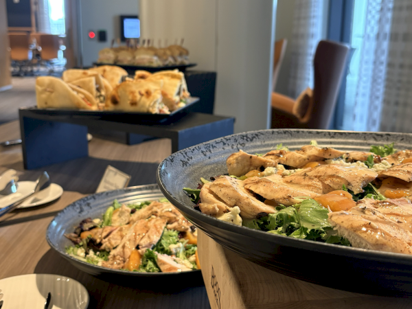 A buffet spread with large bowls of salad and sliced chicken in a bright dining room, set on an outdoor-style serving table.