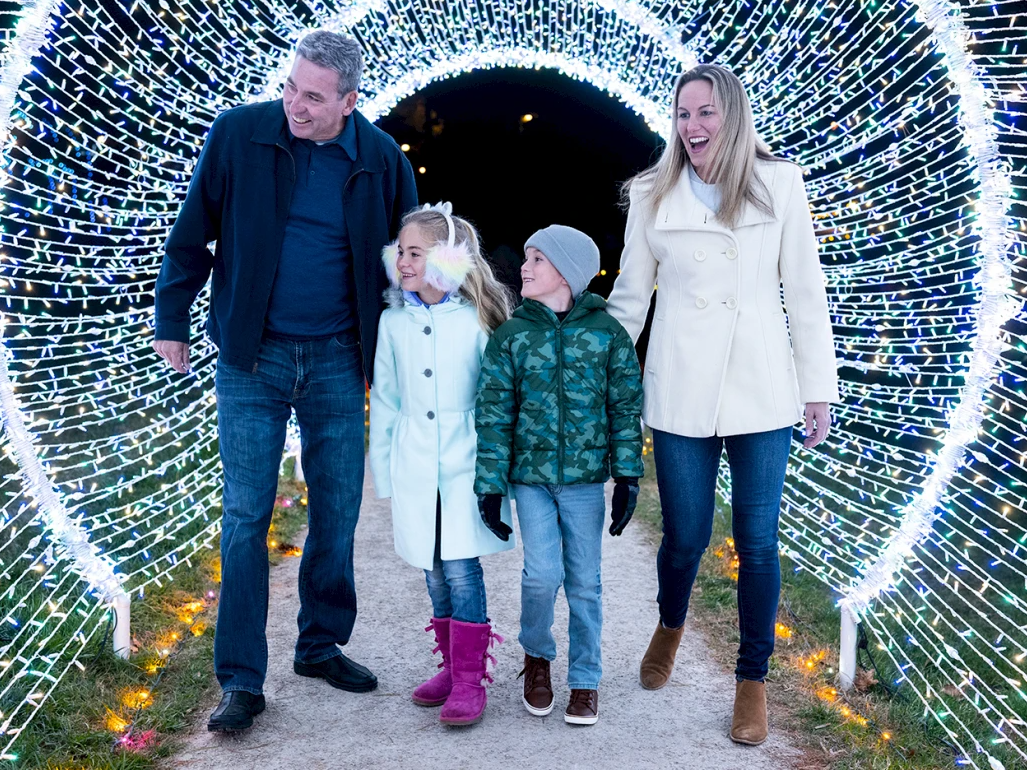 A family of four strolls through a glowing light tunnel, smiling and laughing on a festive winter path.