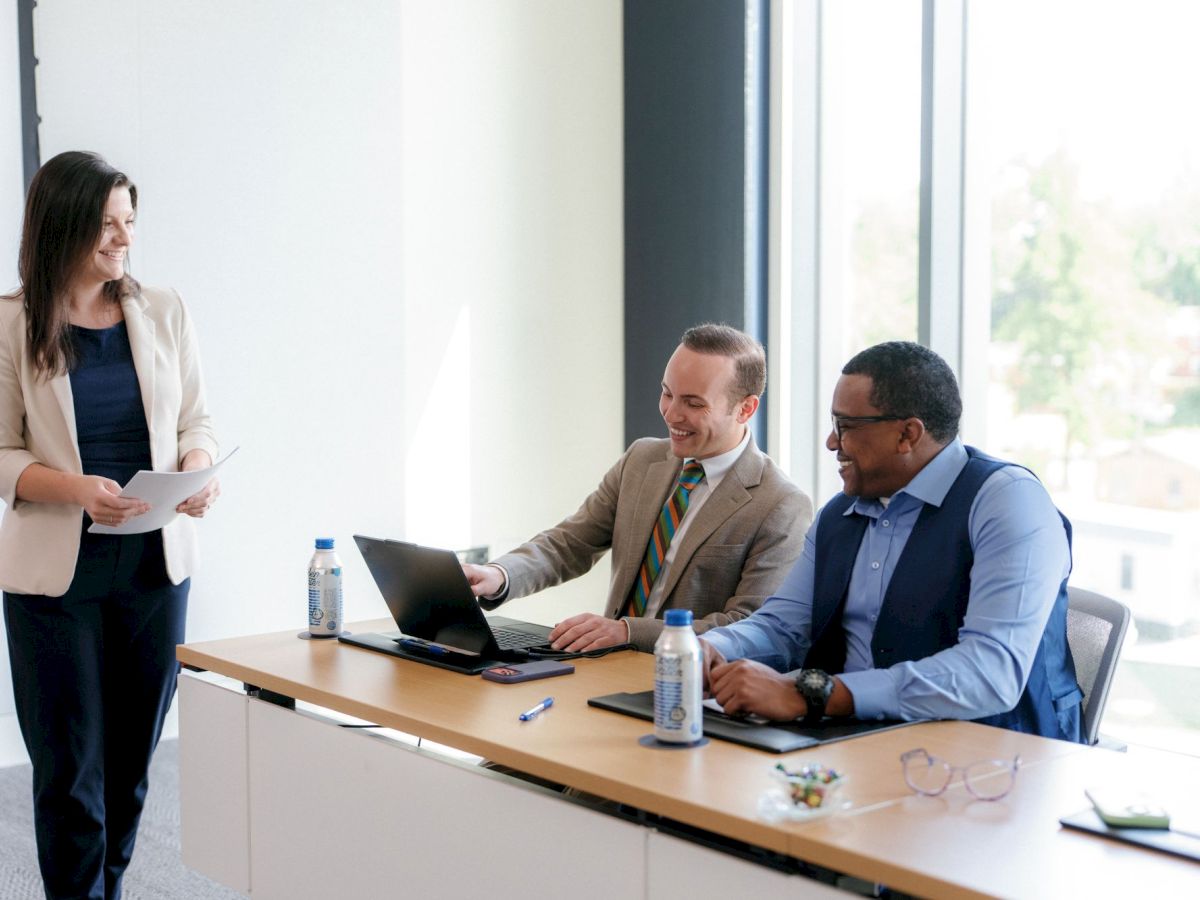 In a bright office, a woman presents papers while coworkers smile and work on laptops during a meeting.